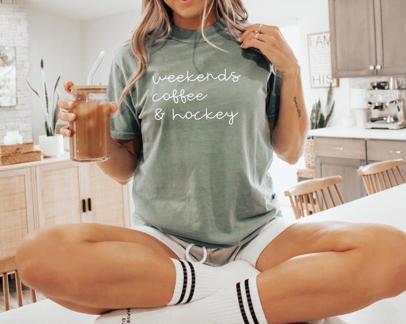 May include: Sage green t-shirt with the words "weekends coffee & hockey" in white script. The person is holding a glass jar with a straw filled with a brown beverage. The person is wearing white shorts and socks with black stripes.