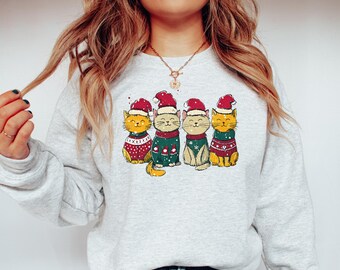 May include: A light gray sweatshirt featuring a graphic of four cartoon cats wearing Santa hats and sweaters. The cats are in various colors, including yellow, cream, and green, with red and white accents. The design has a vintage, slightly distressed look.