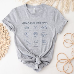 Peut inclure: T-shirt gris avec un motif sur le th&egrave;me du baseball qui dit "NEVER ALLOW THE FEAR OF STRIKING OUT KEEP YOU FROM PLAYING THE GAME." Le motif comprend des illustrations d'&eacute;quipements de baseball, notamment une batte, une balle, un gant, un casque, des crampons et un maillot.