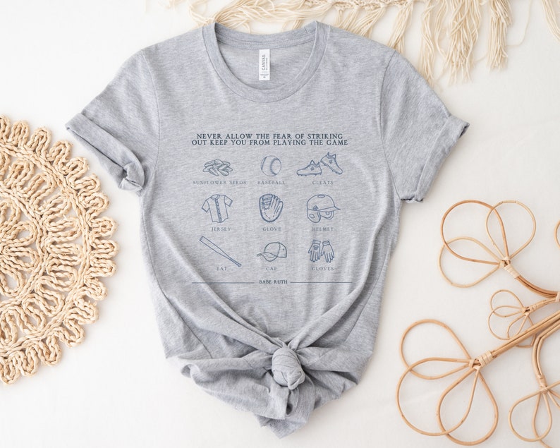 Peut inclure: T-shirt gris avec un motif sur le th&egrave;me du baseball qui dit "NEVER ALLOW THE FEAR OF STRIKING OUT KEEP YOU FROM PLAYING THE GAME." Le motif comprend des illustrations d'&eacute;quipements de baseball, notamment une batte, une balle, un gant, un casque, des crampons et un maillot.