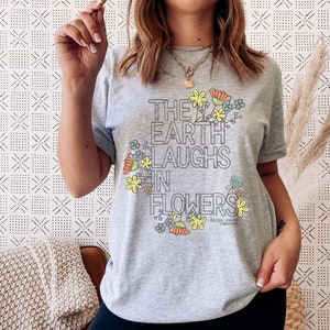 Pode incluir: Camiseta cinza com um design floral e o texto "The Earth Laughs in Flowers" de Ralph Waldo Emerson.