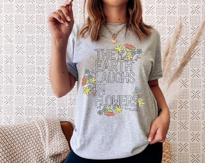 Pode incluir: Camiseta cinza com um design floral e o texto "The Earth Laughs in Flowers" de Ralph Waldo Emerson.