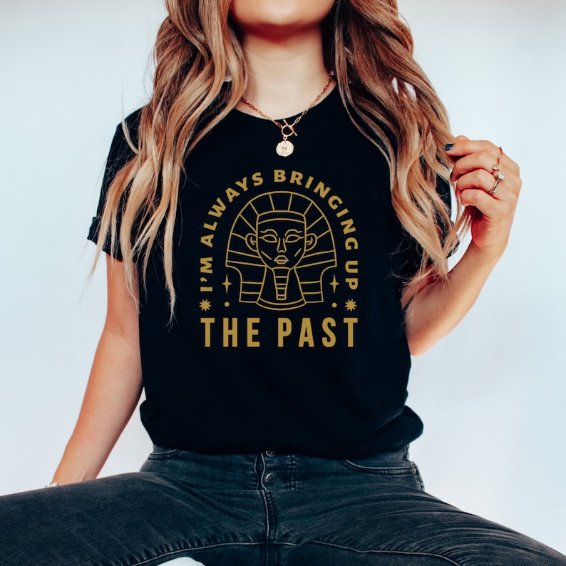 K&ouml;nnte beinhalten: Ein schwarzes T-Shirt mit einer goldenen Grafik eines &auml;gyptischen Pharaonenkopfes in einem Kreis. Der Text "I'm always bringing up the past" ist um den Kreis herum geschrieben.