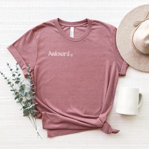 May include: A mauve t-shirt with the text "Awkward" and a heart symbol printed on the front. The shirt is laid flat on a white textured surface with a hat and a mug beside it.