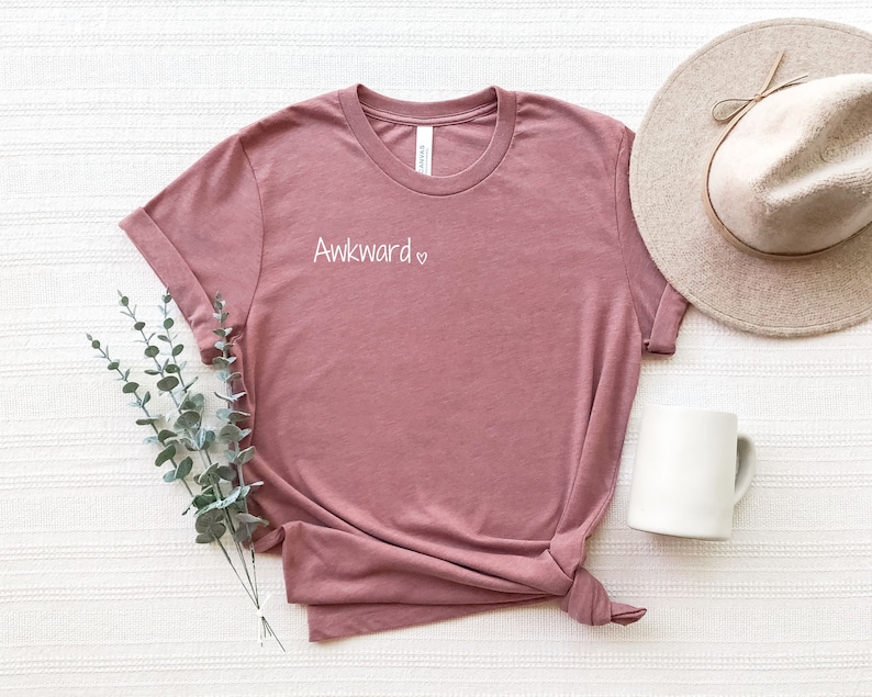 May include: A mauve t-shirt with the text "Awkward" and a heart symbol printed on the front. The shirt is laid flat on a white textured surface with a hat and a mug beside it.