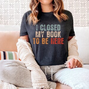 Puede incluir: Una camiseta gris oscuro con un gr&aacute;fico de estilo vintage que dice "Cerr&eacute; mi libro para estar aqu&iacute;".