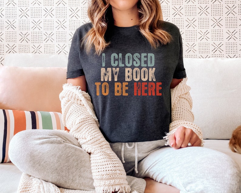 Puede incluir: Una camiseta gris oscuro con un gr&aacute;fico de estilo vintage que dice "Cerr&eacute; mi libro para estar aqu&iacute;".