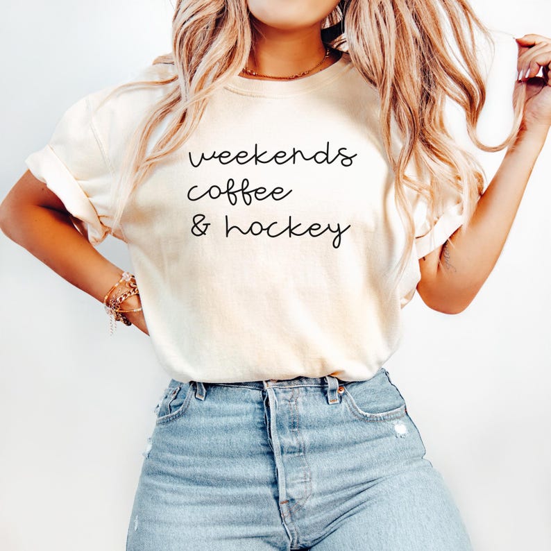 May include: Cream-colored t-shirt with the words "weekends coffee & hockey" in a black, handwritten font. The shirt is paired with blue jeans. The sleeves are rolled up.