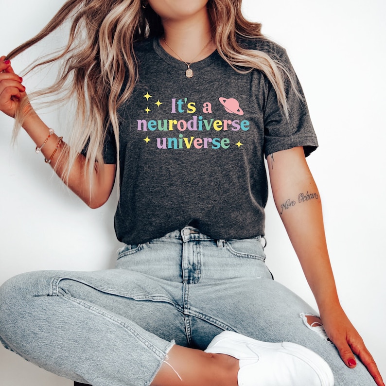 Pu&ograve; includere: Una maglietta grigio scuro con il testo "It's a neurodiverse universe" in colori arcobaleno. Il testo &egrave; circondato da piccole stelle e un pianeta rosa con un anello.