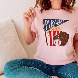 May include: A pink t-shirt with a graphic of a baseball, a glove, popcorn, and a pennant that says "PLAY-BALL".