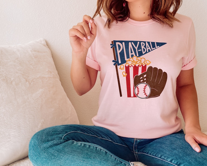 May include: A pink t-shirt with a graphic of a baseball, a glove, popcorn, and a pennant that says "PLAY-BALL".