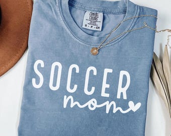 May include: A blue Comfort Colors t-shirt with the words "SOCCER mom" in white script. A gold necklace with a pendant is draped across the shirt. A brown hat is visible in the upper left corner.