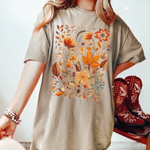 May include: A tan t-shirt featuring a detailed floral design in shades of orange, yellow, and brown. The botanical illustration includes various flowers and leaves. The shirt is a relaxed fit, suitable for casual wear.