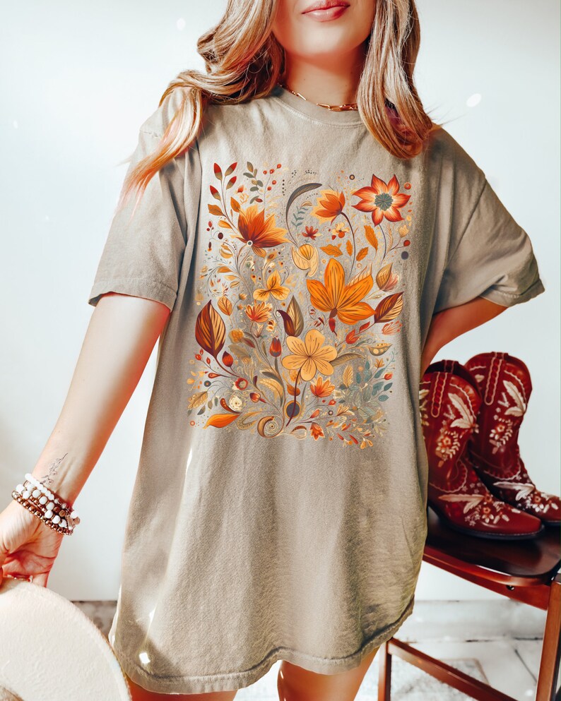May include: A tan t-shirt featuring a detailed floral design in shades of orange, yellow, and brown. The botanical illustration includes various flowers and leaves. The shirt is a relaxed fit, suitable for casual wear.