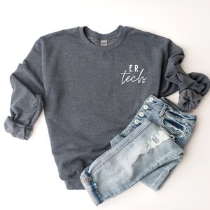 May include: A dark gray sweatshirt with the text "ER tech" in white script on the front. The sweatshirt is paired with light blue denim jeans.