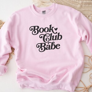 K&ouml;nnte beinhalten: Rosafarbener Pullover mit dem Text "Book Club Babe" in schwarzer Retro-Schrift.