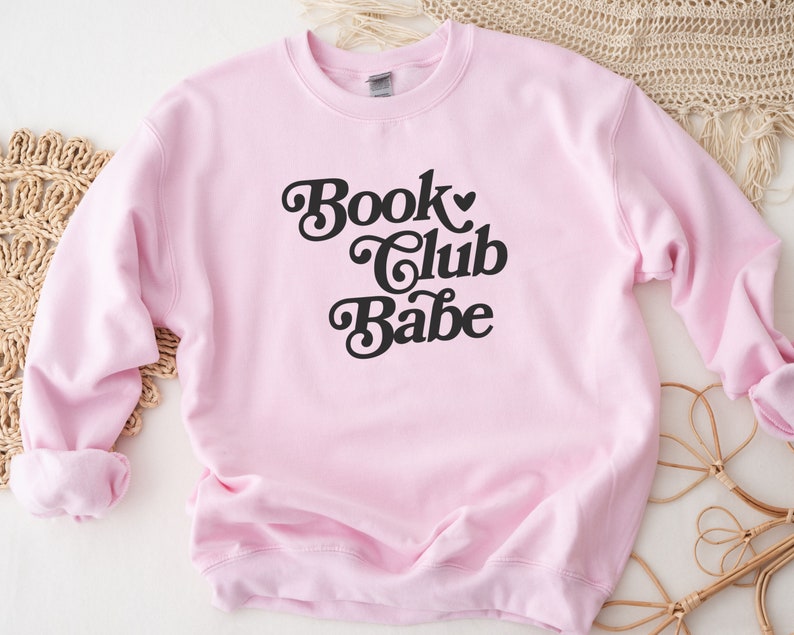 K&ouml;nnte beinhalten: Rosafarbener Pullover mit dem Text "Book Club Babe" in schwarzer Retro-Schrift.