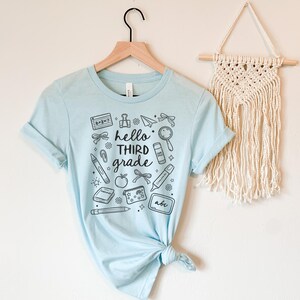 May include: A light blue t-shirt with the words "hello third grade" and school-themed illustrations in black. The design includes a calculator, pencil, apple, and other school supplies. The shirt is hanging on a wooden hanger.
