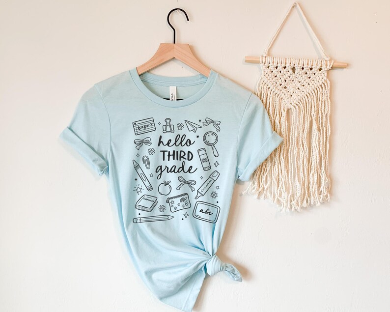 May include: A light blue t-shirt with the words "hello third grade" and school-themed illustrations in black. The design includes a calculator, pencil, apple, and other school supplies. The shirt is hanging on a wooden hanger.