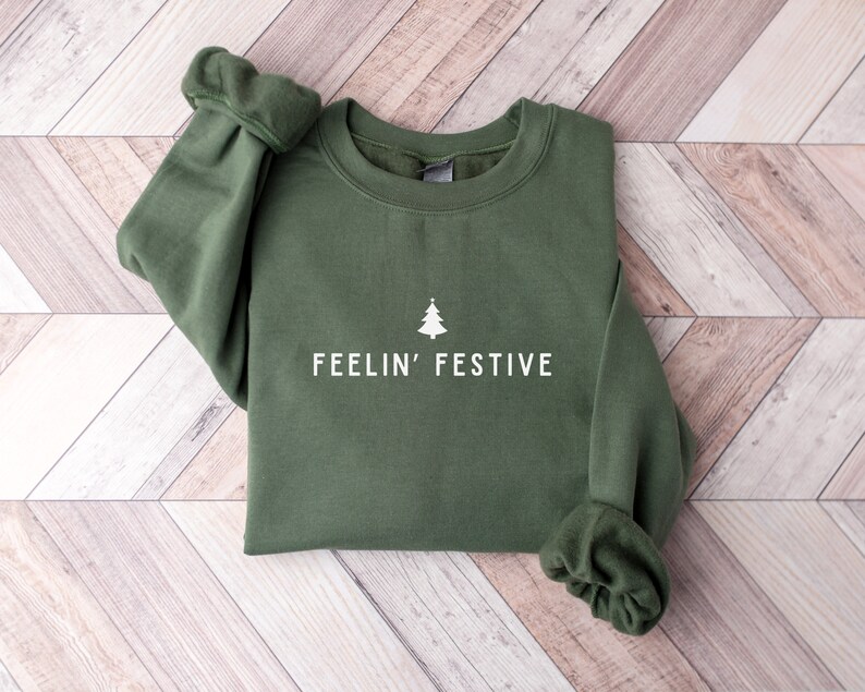 Op de afbeelding: Een groen sweatshirt met een witte kerstboom en de tekst "Feelin' Festive" erop gedrukt.