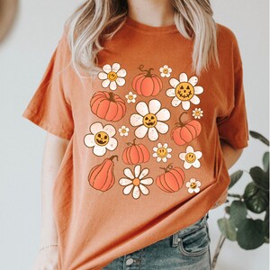 K&ouml;nnte beinhalten: Gebrannt orangefarbenes T-Shirt mit Retro-Design mit orangefarbenen K&uuml;rbissen und wei&szlig;en G&auml;nsebl&uuml;mchen mit l&auml;chelnden Gesichtern. Ein bequemer, l&auml;ssiger Stil, perfekt f&uuml;r Herbst und Halloween.