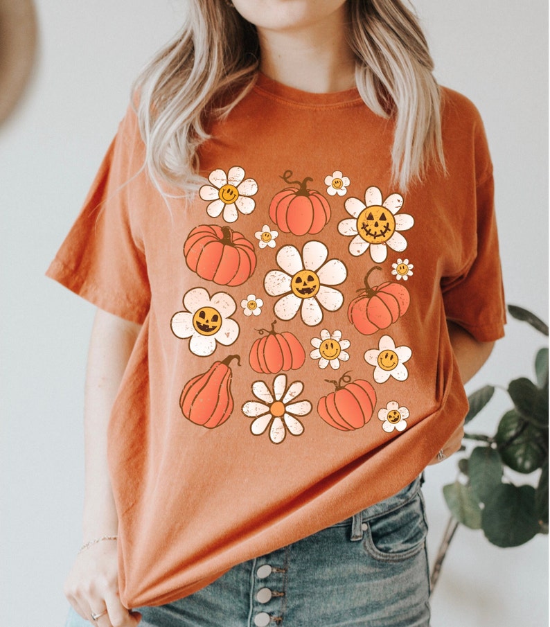 K&ouml;nnte beinhalten: Gebrannt orangefarbenes T-Shirt mit Retro-Design mit orangefarbenen K&uuml;rbissen und wei&szlig;en G&auml;nsebl&uuml;mchen mit l&auml;chelnden Gesichtern. Ein bequemer, l&auml;ssiger Stil, perfekt f&uuml;r Herbst und Halloween.