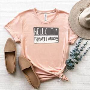 May include: A light pink t-shirt with the text "Hello I'm Probably Anxious" printed on it in a gray box. The shirt is laid out on a white surface with a brown hat and a sprig of eucalyptus.