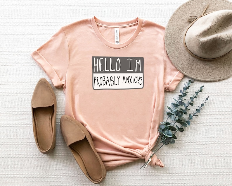 May include: A light pink t-shirt with the text "Hello I'm Probably Anxious" printed on it in a gray box. The shirt is laid out on a white surface with a brown hat and a sprig of eucalyptus.