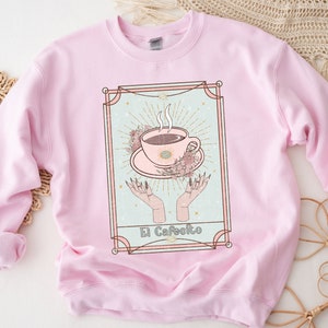 May include: Pink sweatshirt with a graphic of a coffee cup with steam rising from it, surrounded by flowers and hands. The graphic is in a gold frame with the text "El Cafeito" at the bottom.