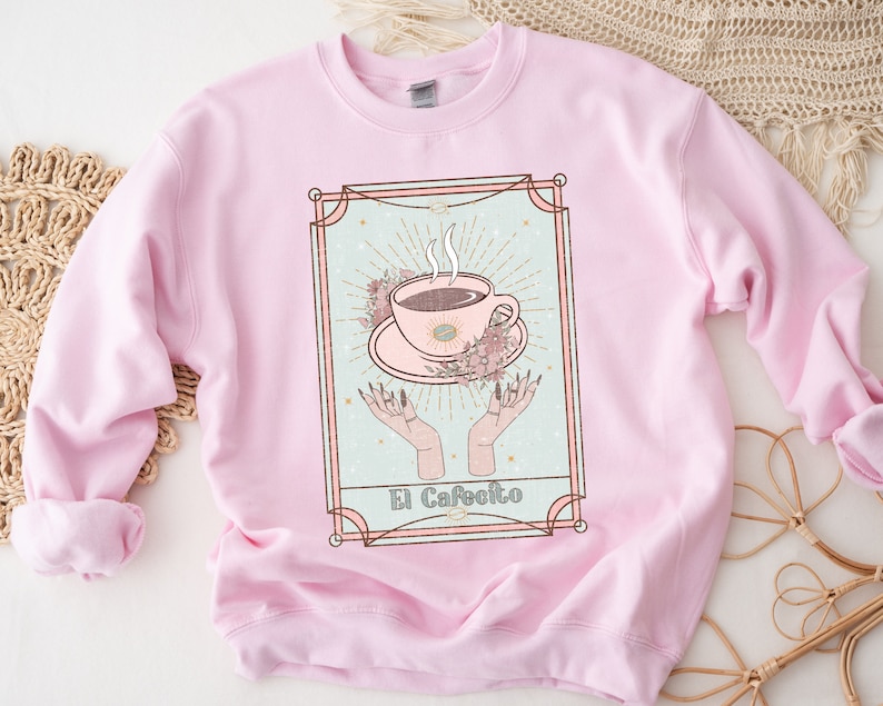 May include: Pink sweatshirt with a graphic of a coffee cup with steam rising from it, surrounded by flowers and hands. The graphic is in a gold frame with the text "El Cafeito" at the bottom.