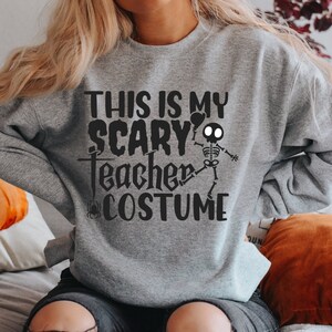 May include: A gray sweatshirt with the text "THIS IS MY SCARY TEACHER COSTUME" in black. A skeleton graphic is on the right side. The sweatshirt is a Halloween-themed garment.