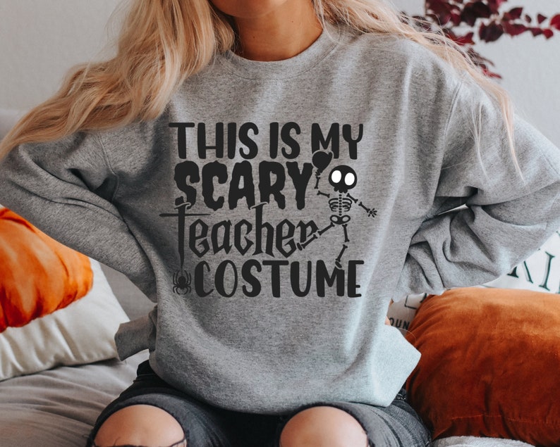 May include: A gray sweatshirt with the text "THIS IS MY SCARY TEACHER COSTUME" in black. A skeleton graphic is on the right side. The sweatshirt is a Halloween-themed garment.