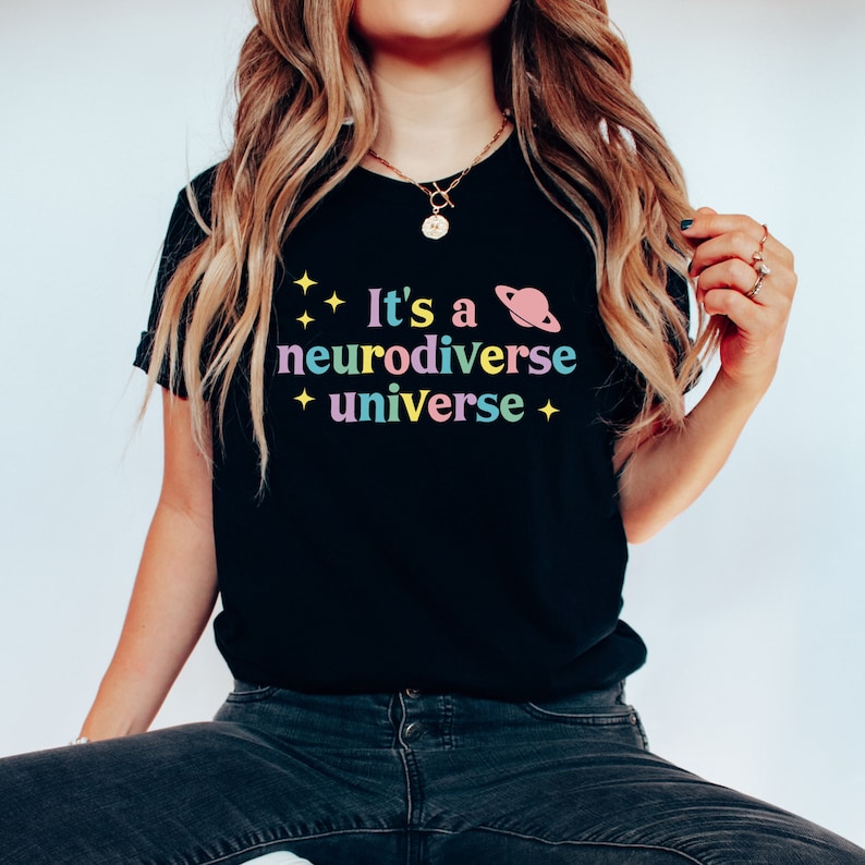 Pu&ograve; includere: Maglietta nera con il testo "It's a neurodiverse universe" in colori arcobaleno con una grafica di un pianeta rosa.