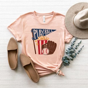 May include: A peach-colored t-shirt with a graphic of a baseball, popcorn, and a glove with the text "Play-Ball" on a pennant.