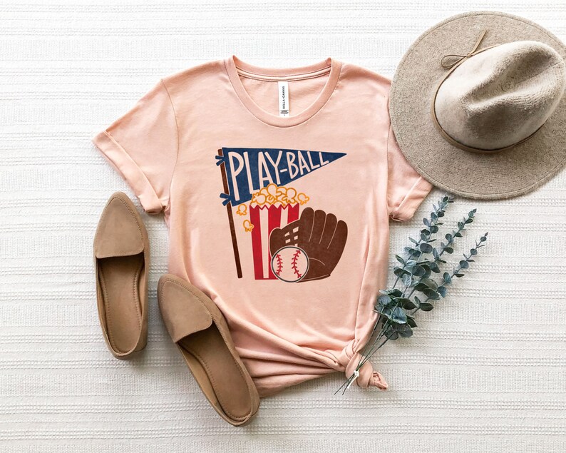 May include: A peach-colored t-shirt with a graphic of a baseball, popcorn, and a glove with the text "Play-Ball" on a pennant.