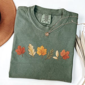 May include: A green t-shirt with a line of fall leaves in orange, yellow, red, and brown. The shirt has the text "COMFORT COLORS" and "S - P - CH" on the front.
