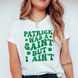 May include: White t-shirt with green text that reads "Patrick was a* Saint *But* I ain't".