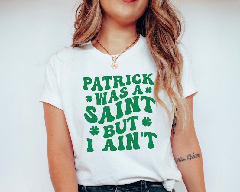 May include: White t-shirt with green text that reads "Patrick was a* Saint *But* I ain't".