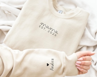 May include: A beige crewneck sweatshirt featuring black text on the chest that reads mama EST. 2026. The sleeve is embroidered with a small black heart icon followed by the name Analise in a simple black script font.