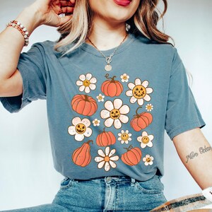 K&ouml;nnte beinhalten: Blau-graues T-Shirt mit einem Retro-Design aus orangefarbenen K&uuml;rbissen und wei&szlig;en G&auml;nsebl&uuml;mchen mit l&auml;chelnden K&uuml;rbisgesichtern. Das Shirt hat einen Vintage-Look, leicht verblichen, perfekt f&uuml;r Herbst oder Halloween.
