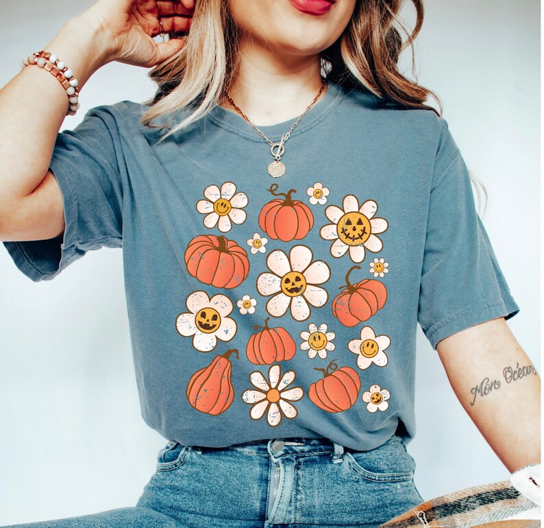 K&ouml;nnte beinhalten: Blau-graues T-Shirt mit einem Retro-Design aus orangefarbenen K&uuml;rbissen und wei&szlig;en G&auml;nsebl&uuml;mchen mit l&auml;chelnden K&uuml;rbisgesichtern. Das Shirt hat einen Vintage-Look, leicht verblichen, perfekt f&uuml;r Herbst oder Halloween.