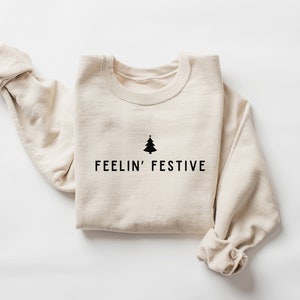 Op de afbeelding: Een beige sweatshirt met een zwarte kerstboom afbeelding en de tekst "FEELIN' FESTIVE".