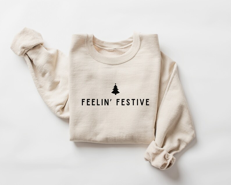 Op de afbeelding: Een beige sweatshirt met een zwarte kerstboom afbeelding en de tekst "FEELIN' FESTIVE".