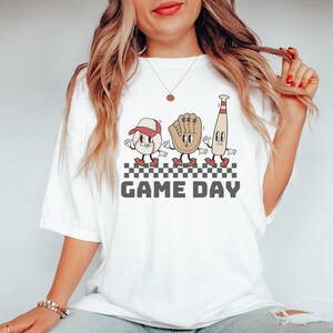 May include: White t-shirt featuring a graphic of a baseball, glove, and bat with cartoon faces. The graphic is on a checkered background with the text "GAME DAY" below it.