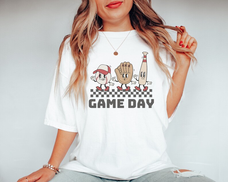 May include: White t-shirt featuring a graphic of a baseball, glove, and bat with cartoon faces. The graphic is on a checkered background with the text "GAME DAY" below it.