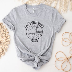 Puede incluir: Camiseta gris con una ilustraci&oacute;n en blanco y negro de un faro y el texto "Cabot Cove, Maine Est 1984".