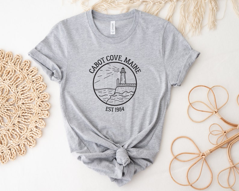 Puede incluir: Camiseta gris con una ilustraci&oacute;n en blanco y negro de un faro y el texto "Cabot Cove, Maine Est 1984".