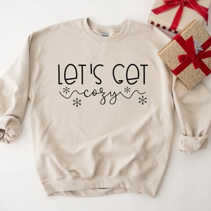 May include: A beige sweatshirt with the text "Let's Get Cozy" in black lettering with snowflakes on either side.
