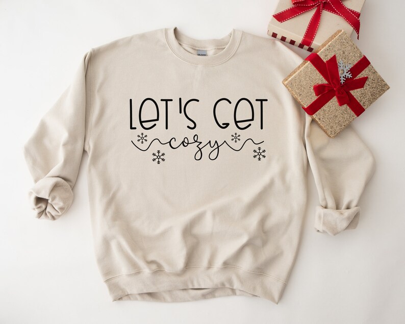 May include: A beige sweatshirt with the text "Let's Get Cozy" in black lettering with snowflakes on either side.