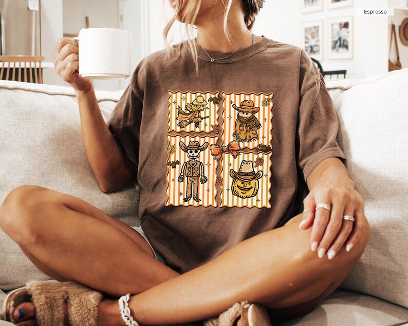 May include: A brown t-shirt with a four-panel Halloween design. The panels feature a witch, scarecrow, skeleton, and pumpkin wearing a cowboy hat. The background has orange and white vertical stripes. The shirt is espresso-colored.